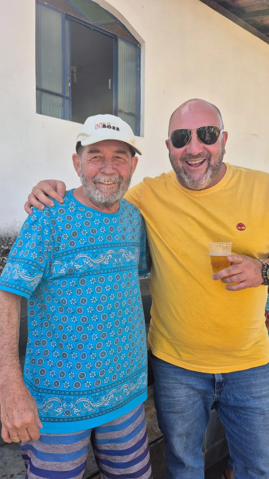 O Senhor Lakes, de boné e camisa azul, abraçado com Warley, de camiseta amarela, em frente ao bar.